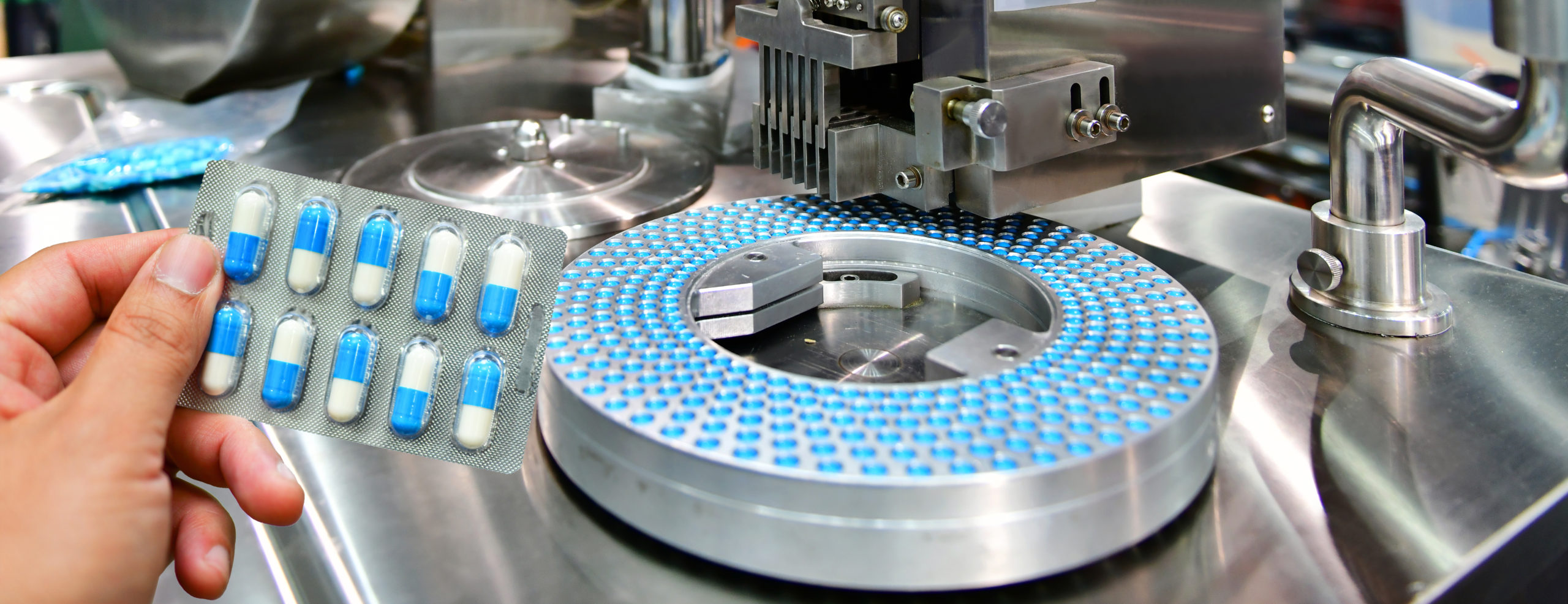 Medicines manufacturing production line, with hand holding pack of blue capsules in foreground